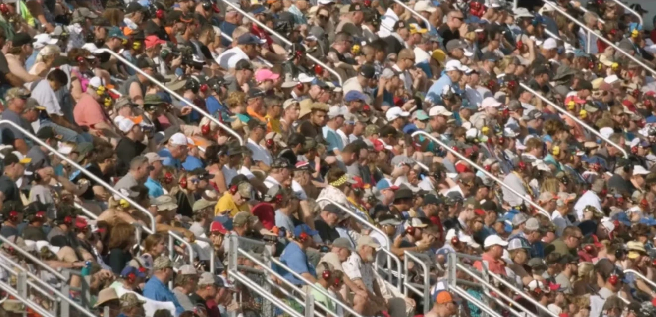 Large crowd of spectators fill stadium seating, enjoying an outdoor event with a mix of hats, shirts, and sunlit views, creating a lively atmosphere.