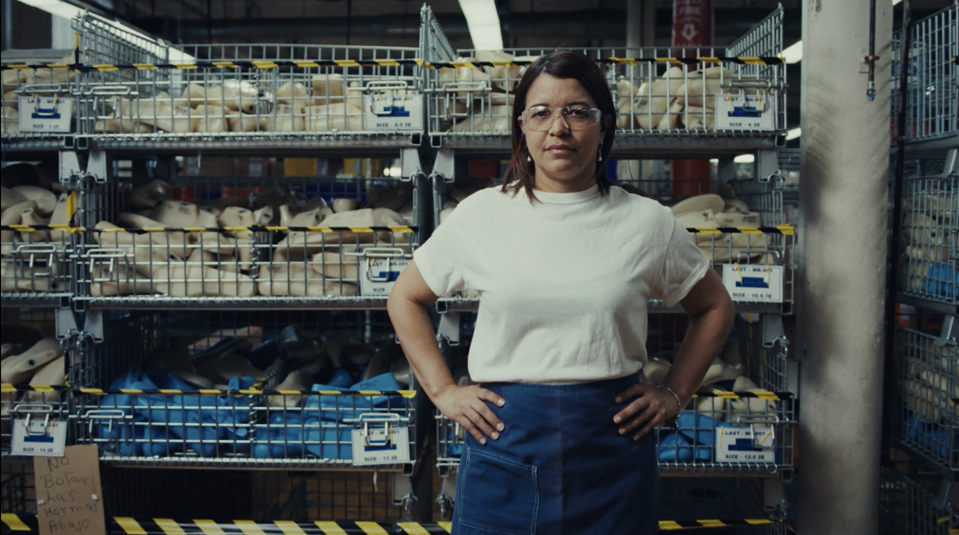 A woman with glasses and a white t-shirt stands in a warehouse with racks of shoe lasts in baskets, creating a serious but neutral mood.