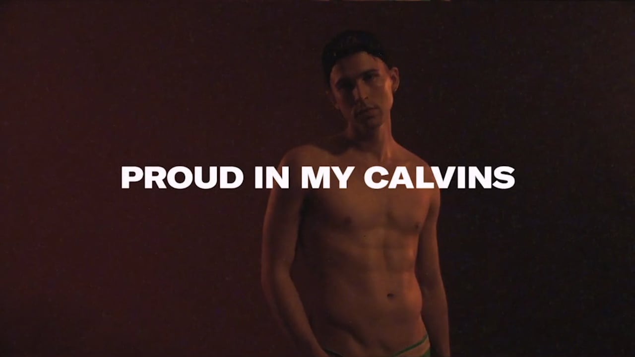Shirtless adult male model poses with a dark backdrop in a medium shot, showcasing his physique with 'Proud in my Calvins' text overlay.