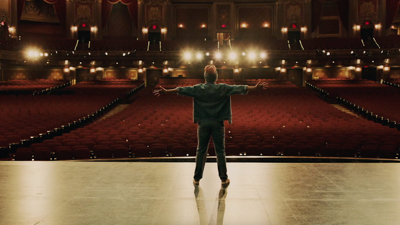Solo performer stands center stage, arms outstretched, in a grand theater with red velvet seating and ornate gold detailing.
