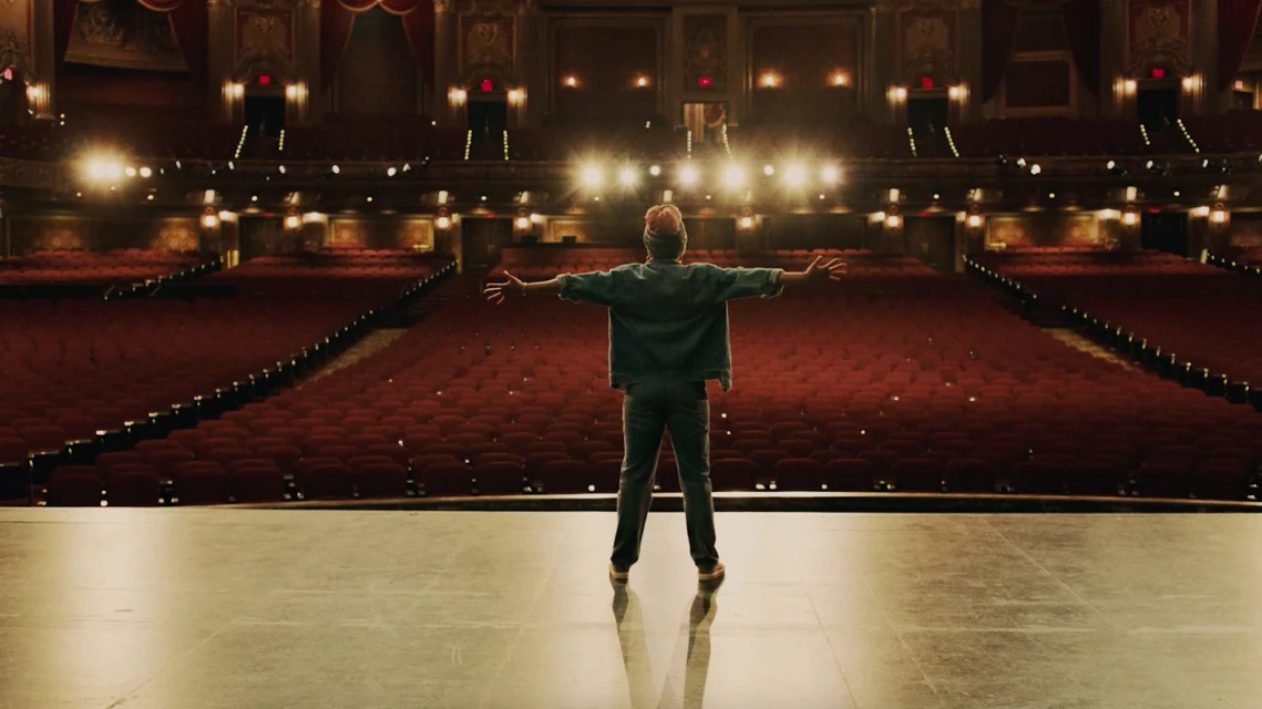 Solo performer stands center stage, arms outstretched, in a grand theater with red velvet seating and ornate gold detailing.