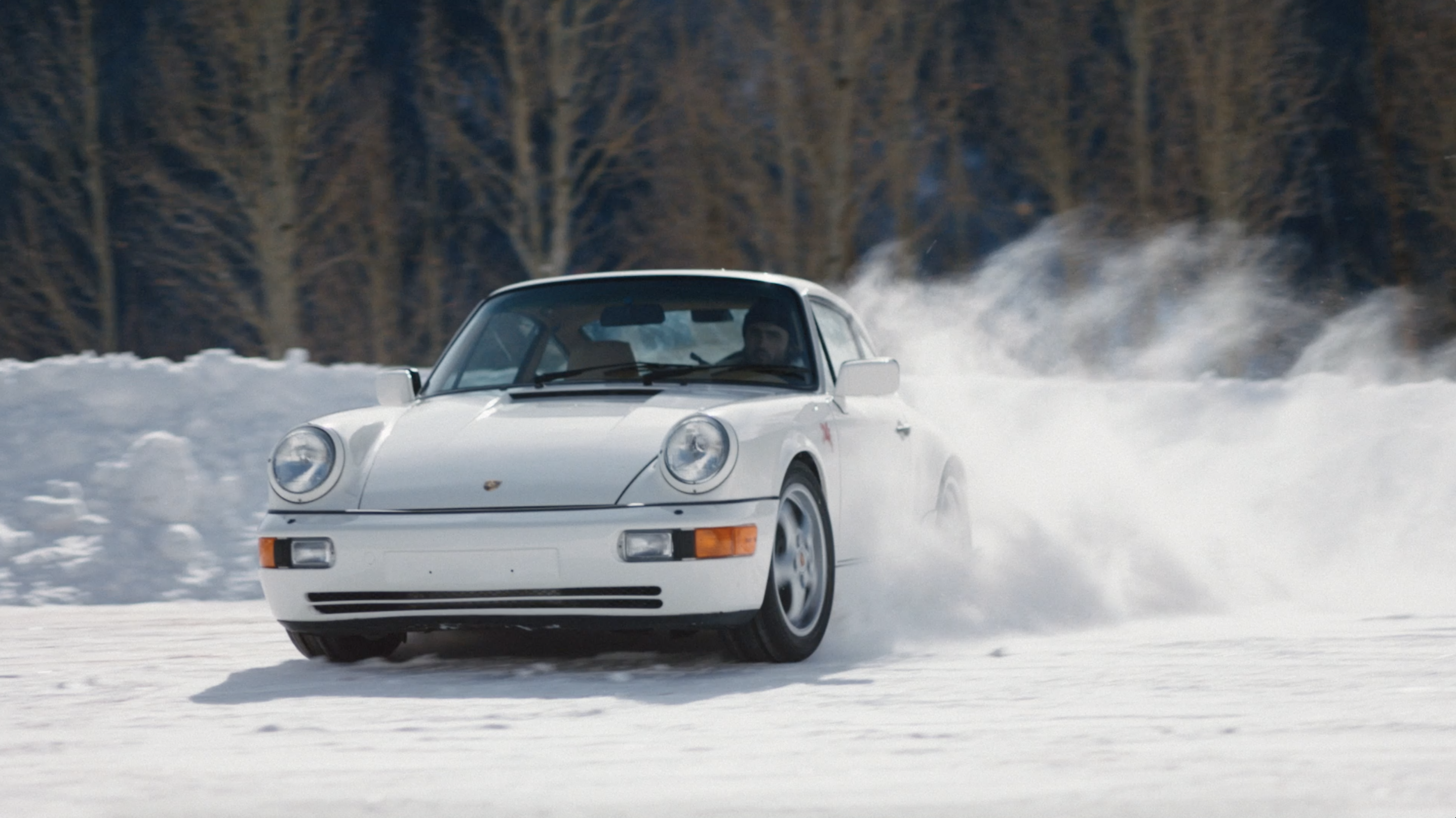 White Porsche 911 drifts on a snowy surface with bare trees visible in the background, capturing a sense of winter sports and automotive.