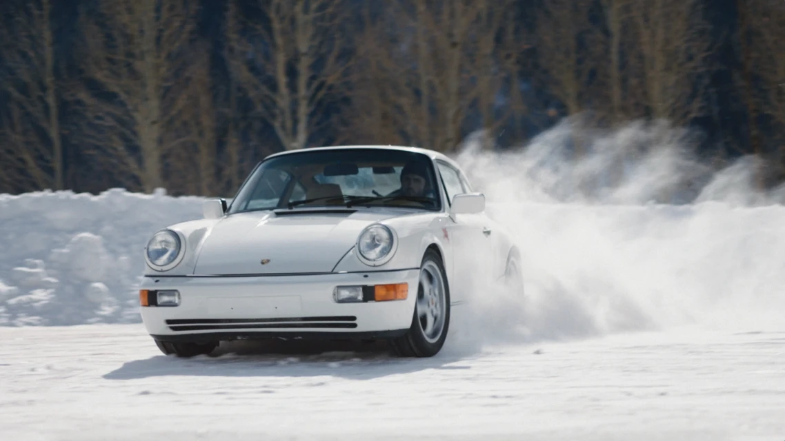 White Porsche 911 drifts on a snowy surface with bare trees visible in the background, capturing a sense of winter sports and automotive.