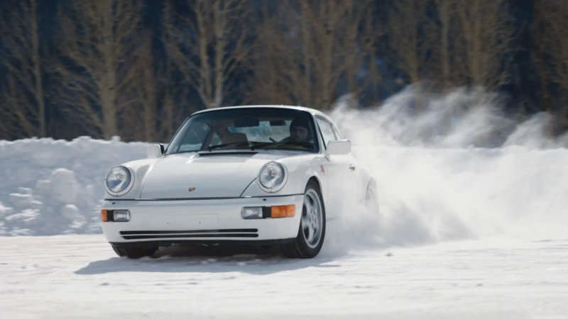 White Porsche 911 drifts on a snowy surface with bare trees visible in the background, capturing a sense of winter sports and automotive.