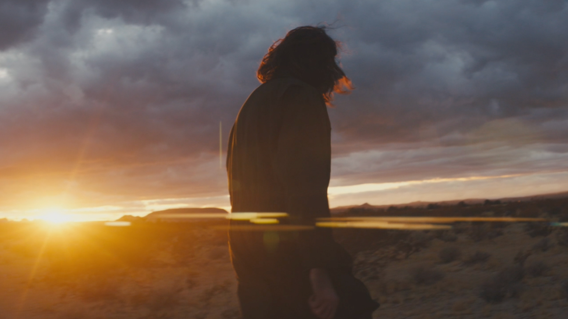 Adult wearing a jacket stands in a desert landscape at sunset, with a dramatic sky of dark clouds tinged with warm sunset colors.
