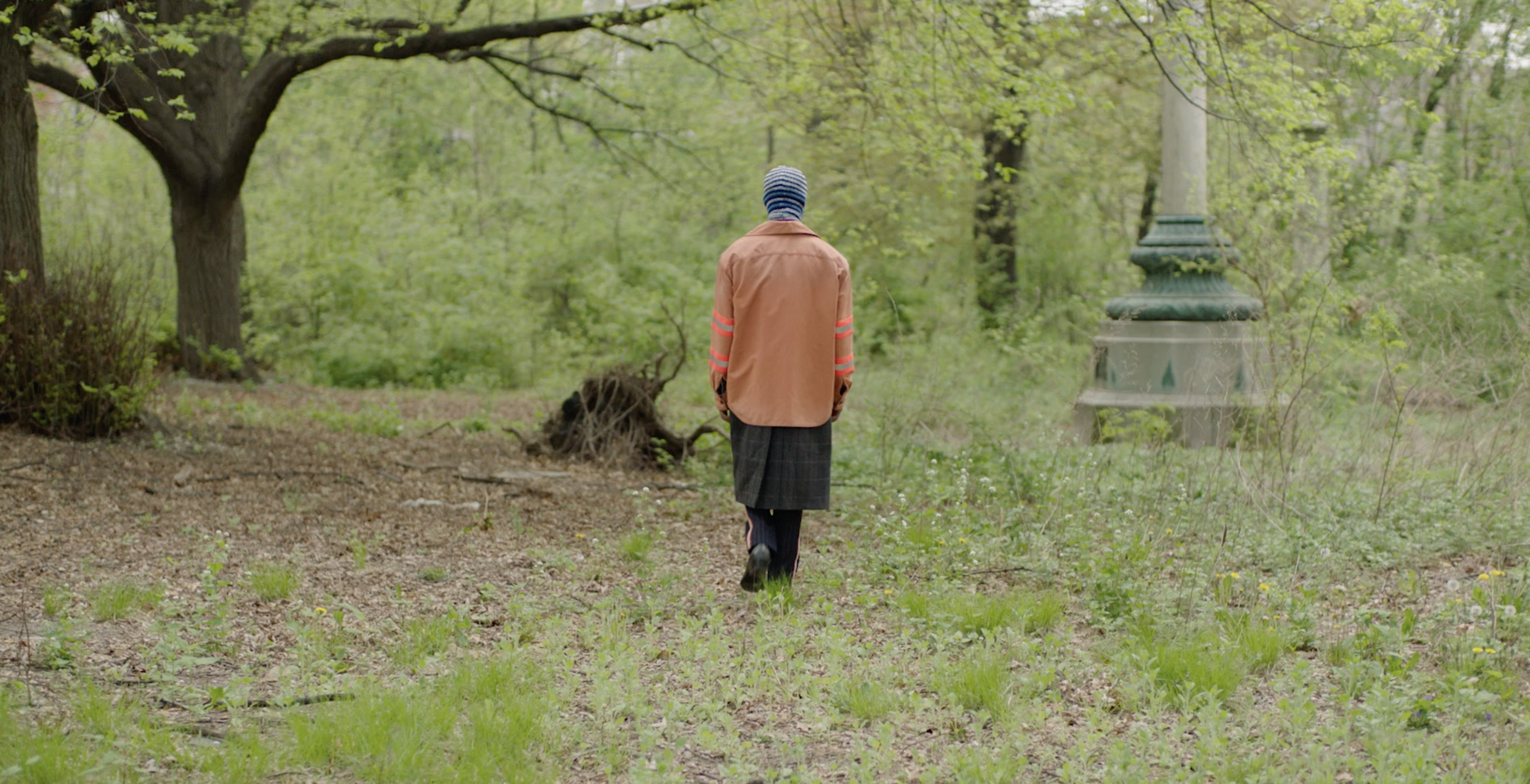 A person walks through a park-like setting wearing an orange shirt, gray skirt, and striped beanie hat, surrounded by trees and greenery.