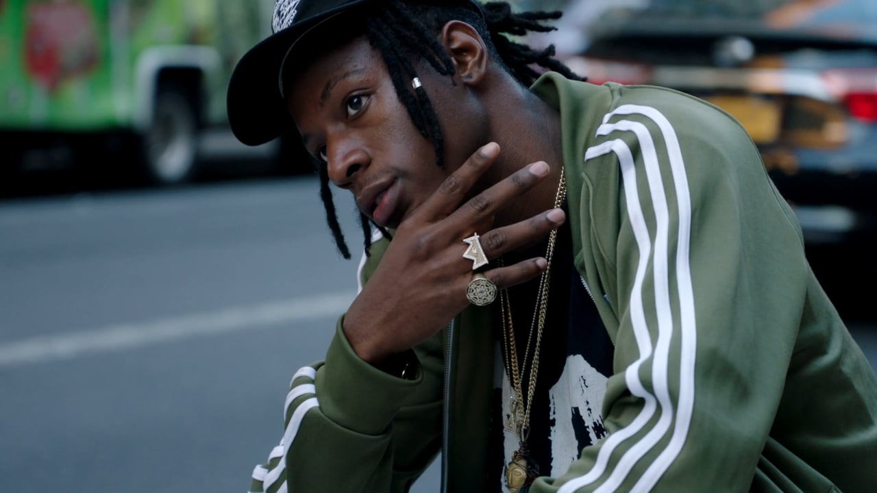 Adult male with dreadlocks, wearing a jacket and gold chains, posing outdoors in daylight, with a blurred car in the background.