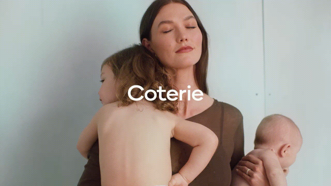 Woman poses with two children in front of a pale blue wall. She wears a brown top, while the kids are bare-chested, with the word 'Coterie'.