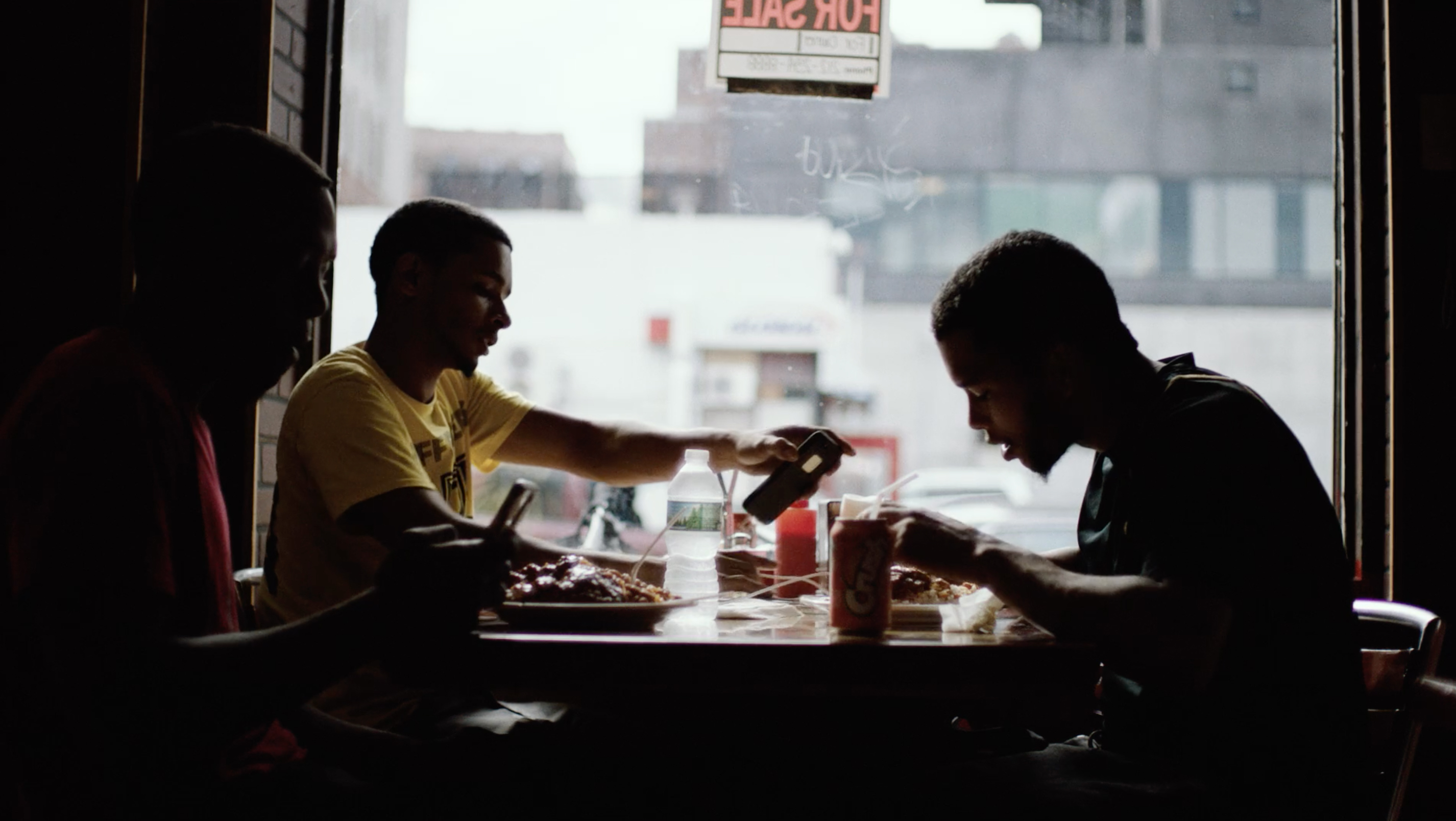 Three men of African descent are silhouetted in a restaurant eating food at a table, with a window and city street in the background.