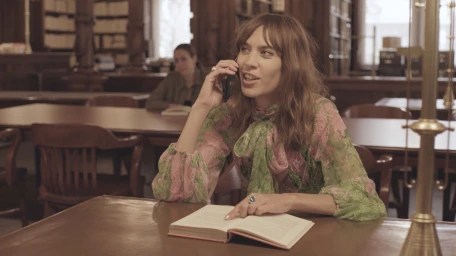 Woman in pink and green patterned blouse talking on a smartphone inside a library with wooden desks
