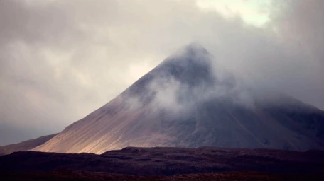 A massive volcanic mountain peak partially shrouded in low-hanging clouds and mist under an overcast sky.