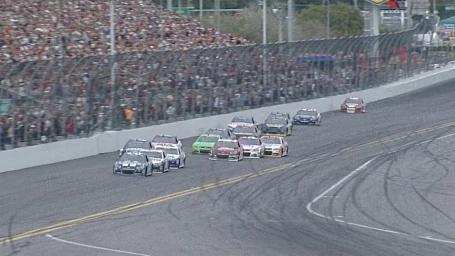 Multiple stock cars racing on an asphalt speedway curve with packed grandstands full of spectators in the background.