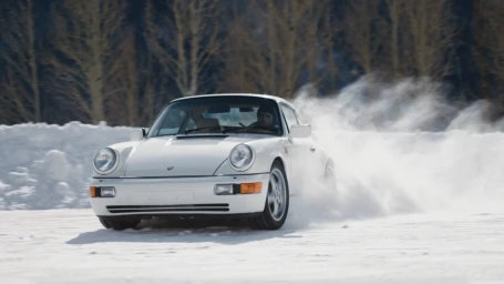A white sports car drifting on a snow-covered track, kicking up a large cloud of powder snow in a wintry landscape.
