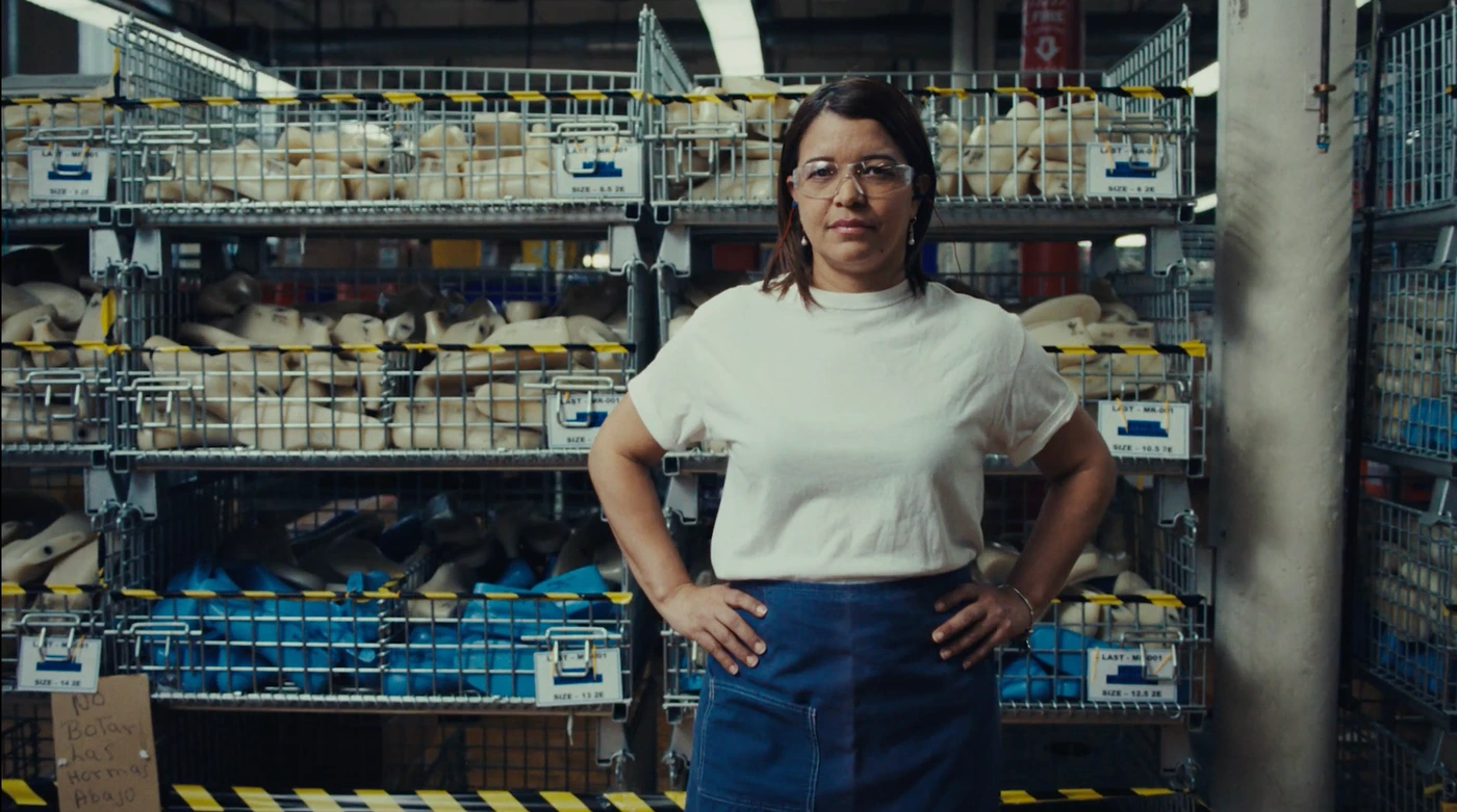 A woman wearing safety glasses, a white t-shirt, and a blue apron stands with hands on hips inside a wire-cage warehouse facility.