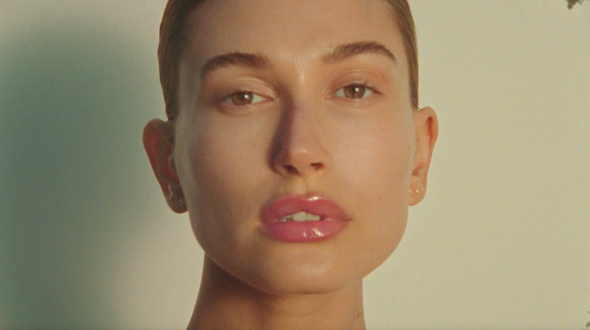 Close up portrait of Hailey Bieber with glossy lips and minimal makeup against a neutral gradient background