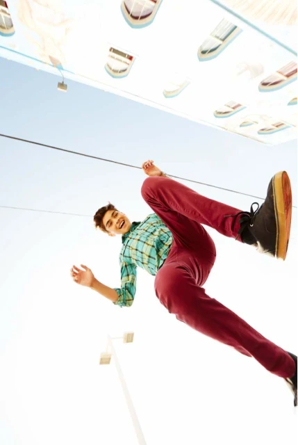 A young adult male in a blue plaid shirt and maroon pants strikes a dynamic pose with shoes and a light blue sky in the background.
