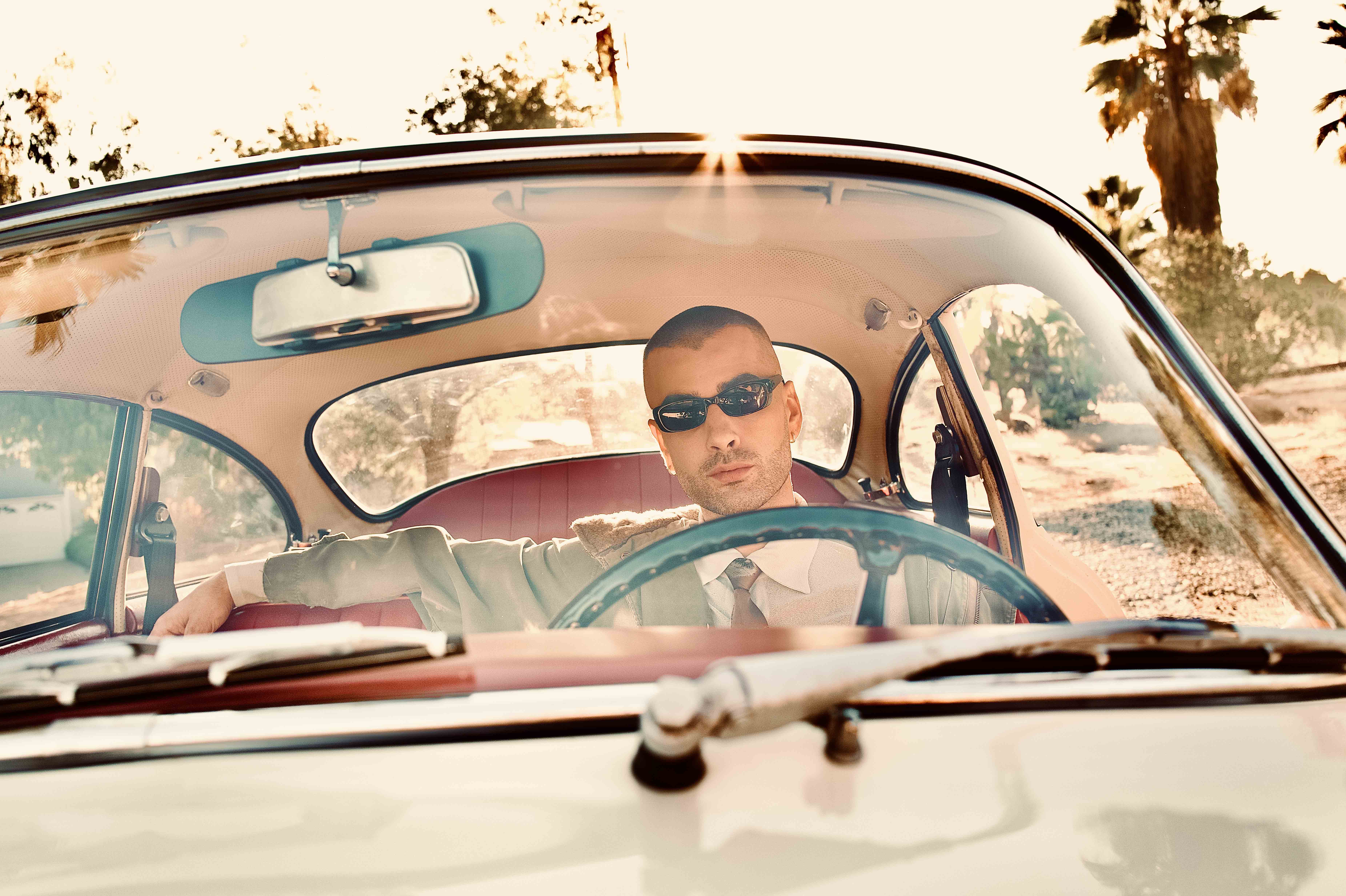 A man with sunglasses drives a classic car in a desert landscape, captured through the windshield in warm, sun-drenched lighting.