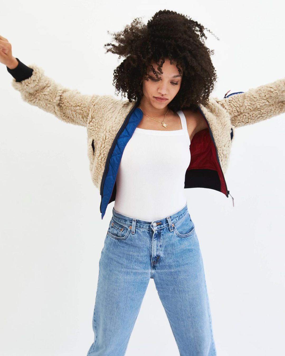 A young woman with curly hair poses in a studio wearing a fuzzy jacket, a white tank top, and light wash jeans against a white background.