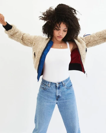 A young woman with curly hair poses in a studio wearing a fuzzy jacket, a white tank top, and light wash jeans against a white background.