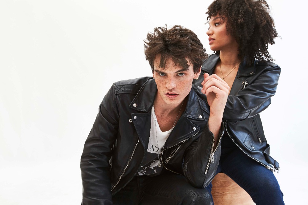 Two fashion models pose together in black leather jackets against a bright white backdrop in a studio setting, showcasing apparel.
