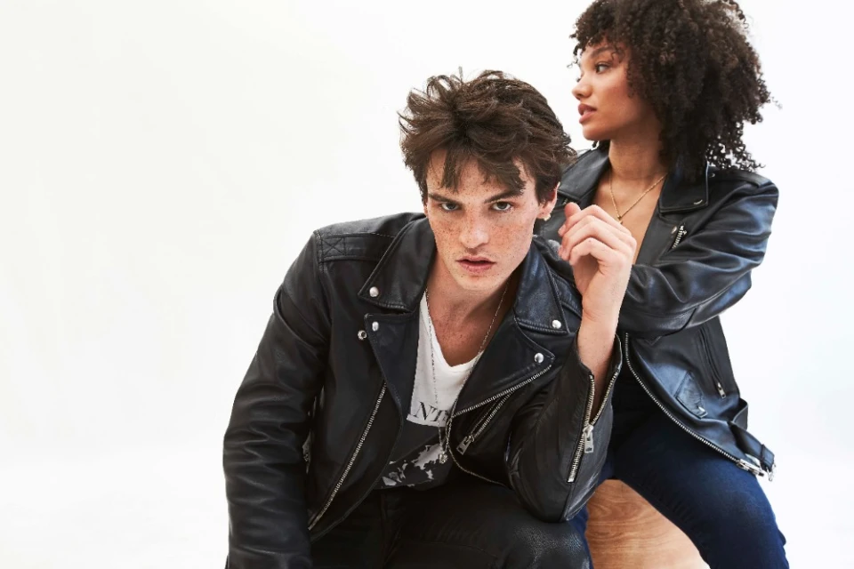 Two fashion models pose together in black leather jackets against a bright white backdrop in a studio setting, showcasing apparel.