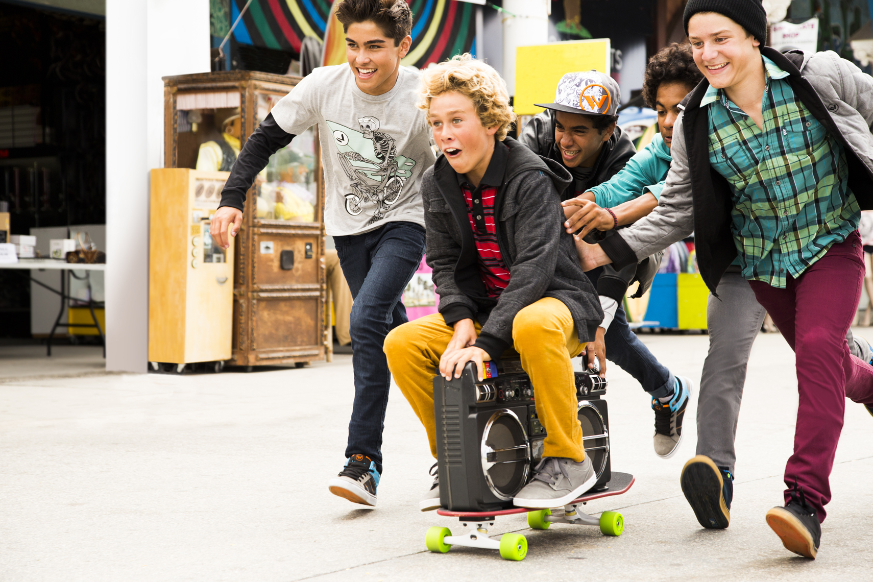 Teenagers push boy on a skateboard with a boombox outdoors in an urban setting, with the group smiling, dressed in casual streetwear.