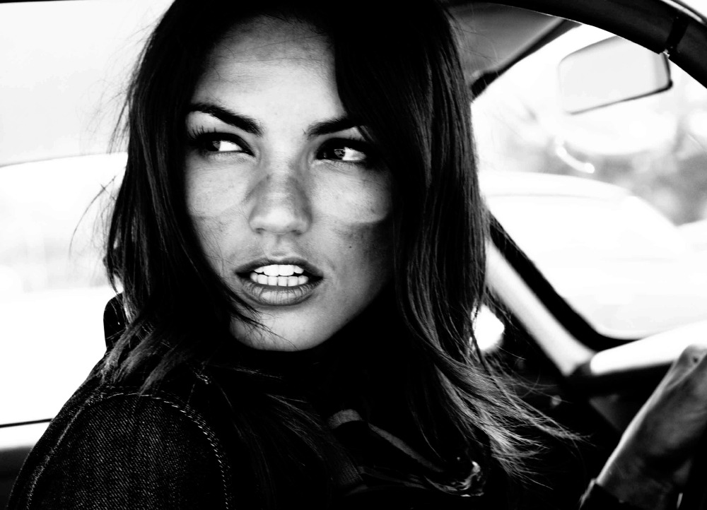 Black and white portrait of a woman driving a car, looking over her shoulder with an open mouth and visible teeth.