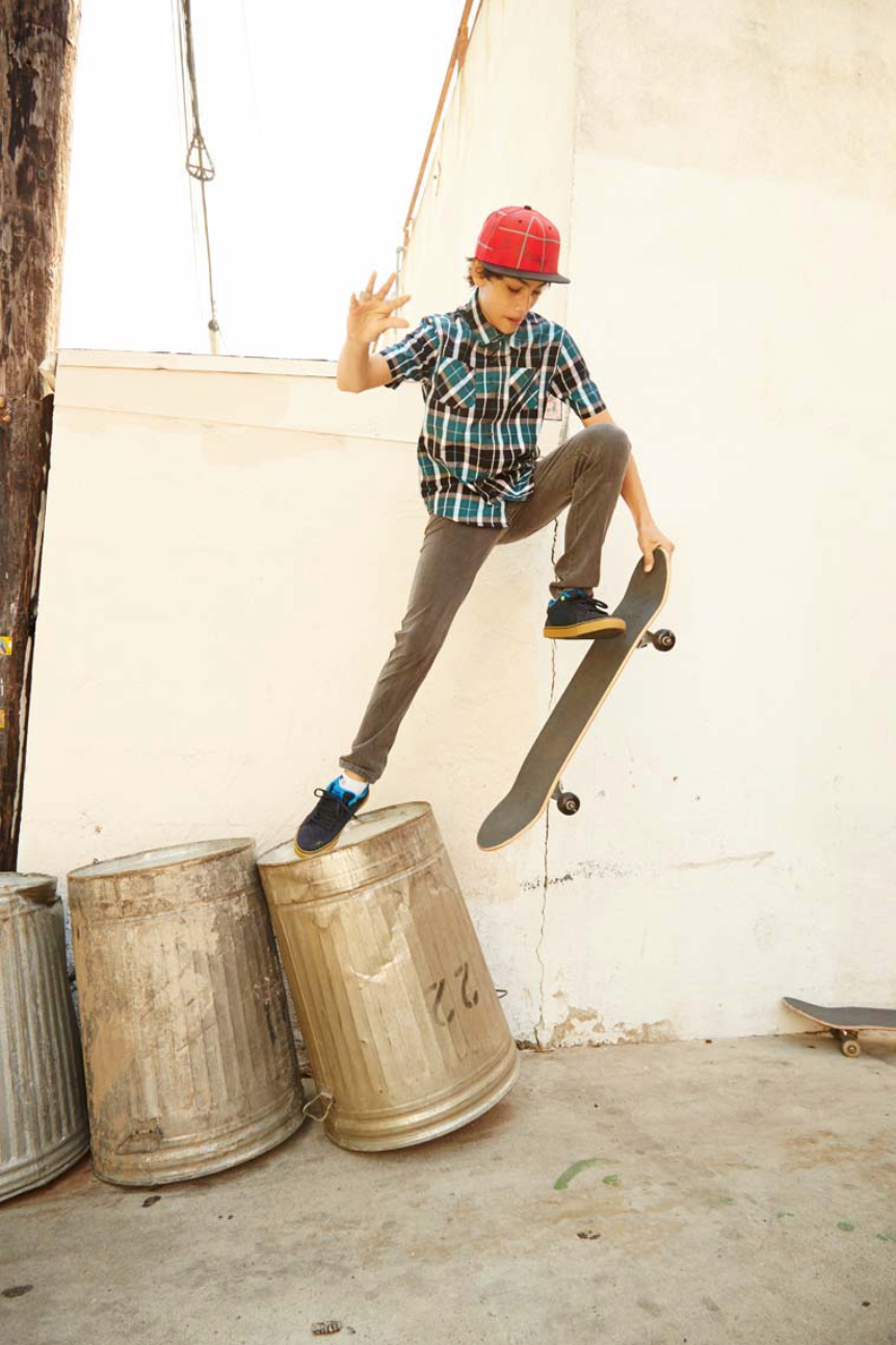 Boy in mid-air, skateboarding over metal trash cans, wearing a red cap and plaid shirt, with a white brick wall background.