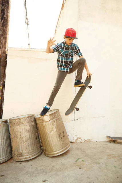 Boy in mid-air, skateboarding over metal trash cans, wearing a red cap and plaid shirt, with a white brick wall background.