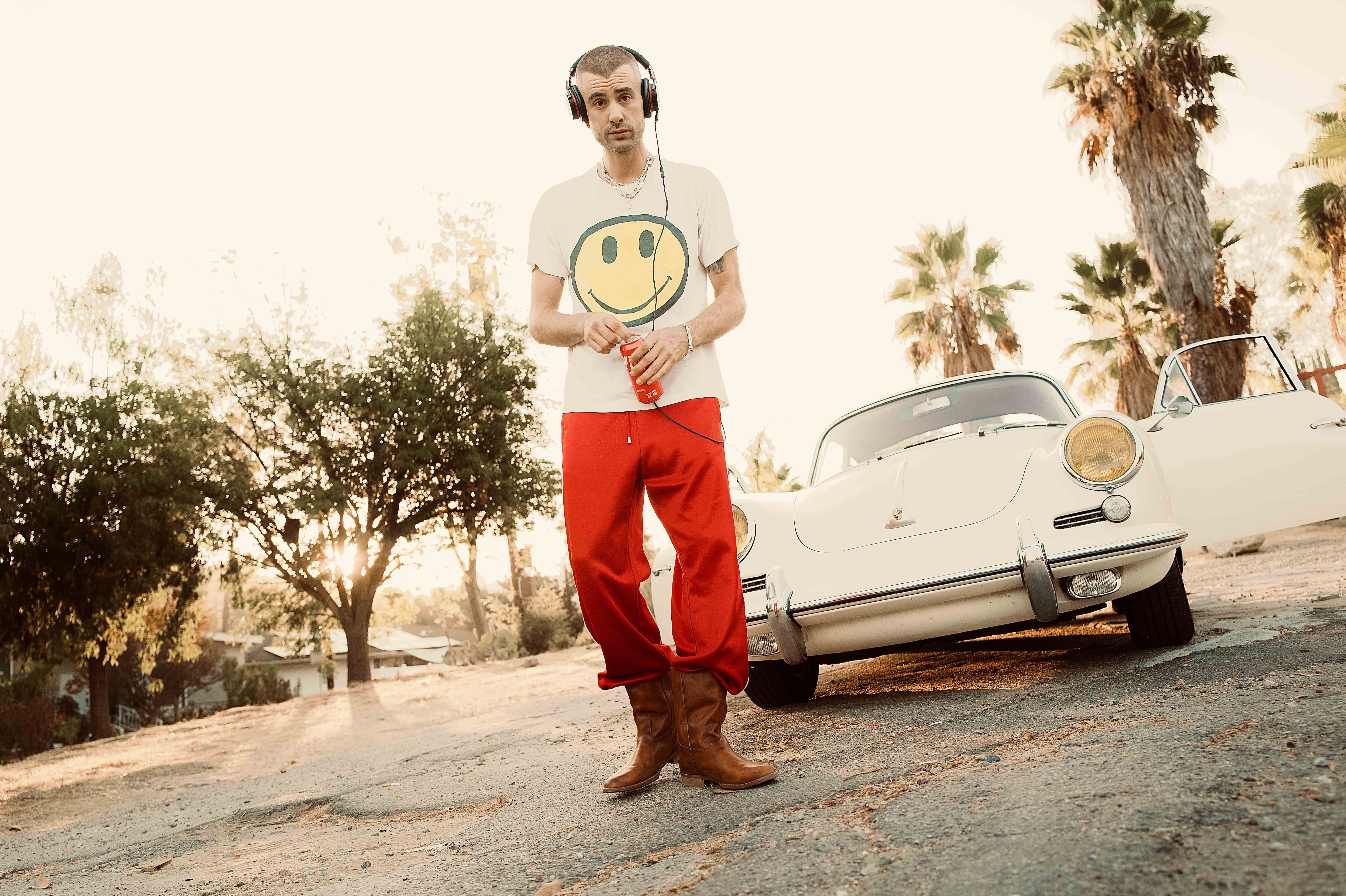 A man stands outside next to a classic car wearing headphones and red pants, with palm trees and blue sky visible in the background.