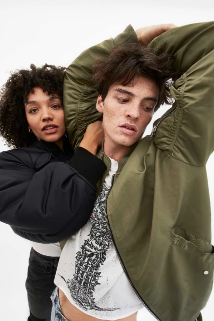 Two adult models, one African-American woman in black jacket and one Caucasian man in olive bomber, pose in front of white backdrop.