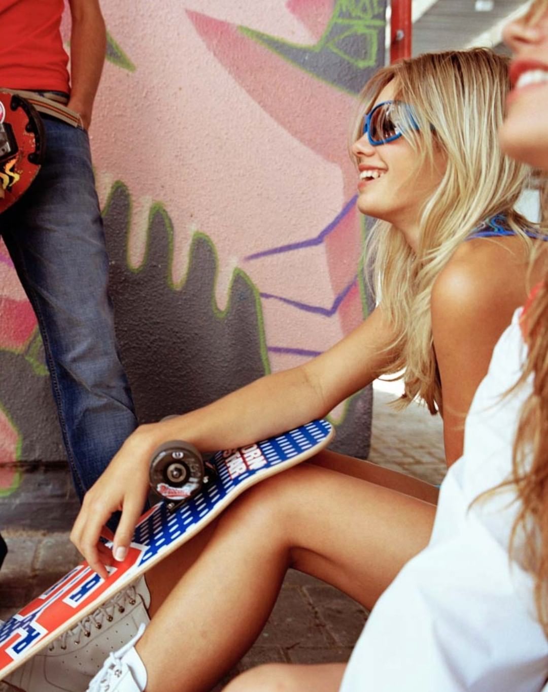 Two young women seated with a skateboard, smiling against graffiti wall, with a young man in the background.