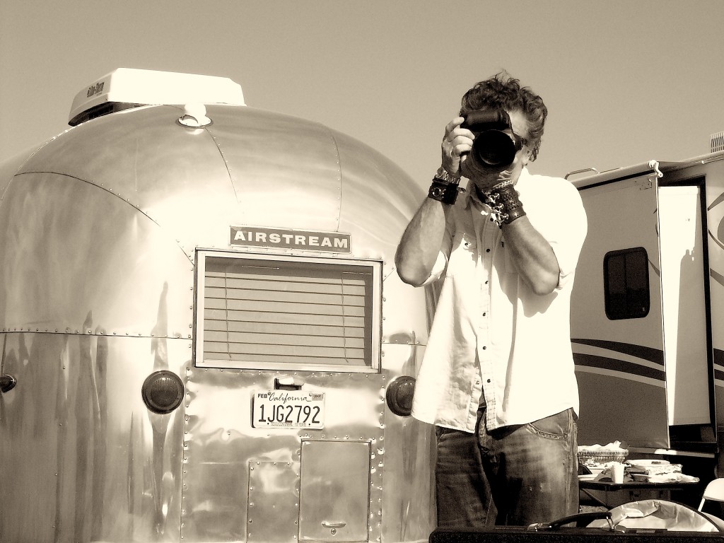 Man in white shirt takes a photo with a DSLR camera in front of a shiny Airstream trailer on a sunny day in a sepia-toned outdoor setting.
