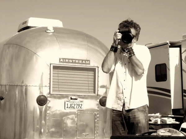 Man in white shirt takes a photo with a DSLR camera in front of a shiny Airstream trailer on a sunny day in a sepia-toned outdoor setting.