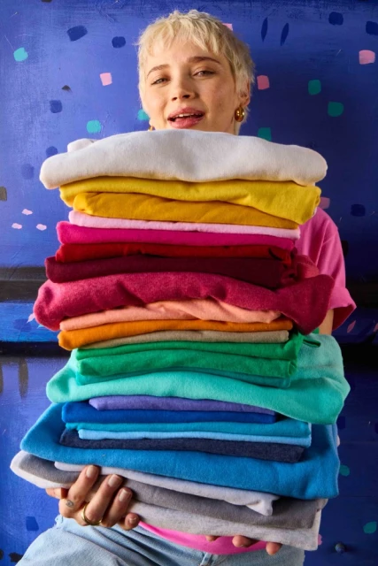 Young woman smiles and holds a tall stack of folded sweatshirts in many colors against a blue abstract background. Colorful clothes.