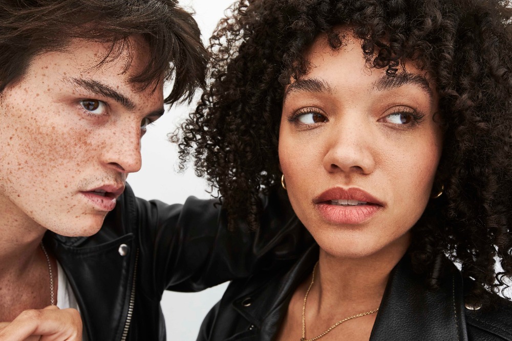 Close-up portrait of two young models wearing black leather jackets against a plain white background, photographed by Larry Bartholomew.