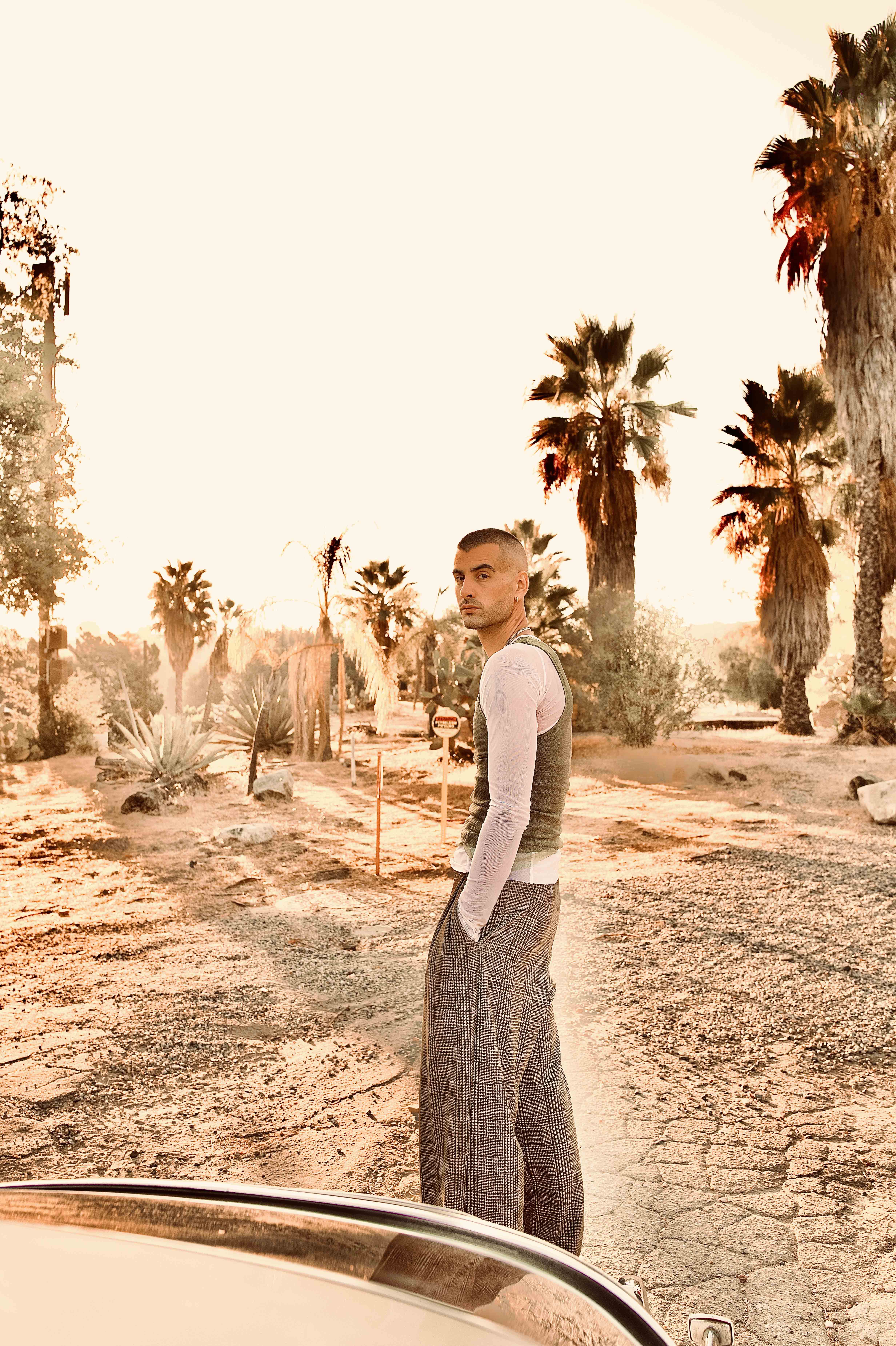 Adult male fashion model poses outdoors in desert setting at dusk. Palm trees in background and part of car visible in foreground.