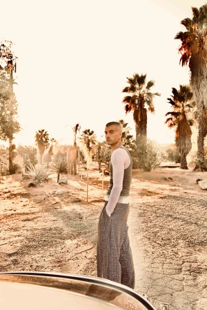 A man with short hair wearing a ribbed green tank top and grey patterned trousers posing outdoors among palm trees in Los Angeles