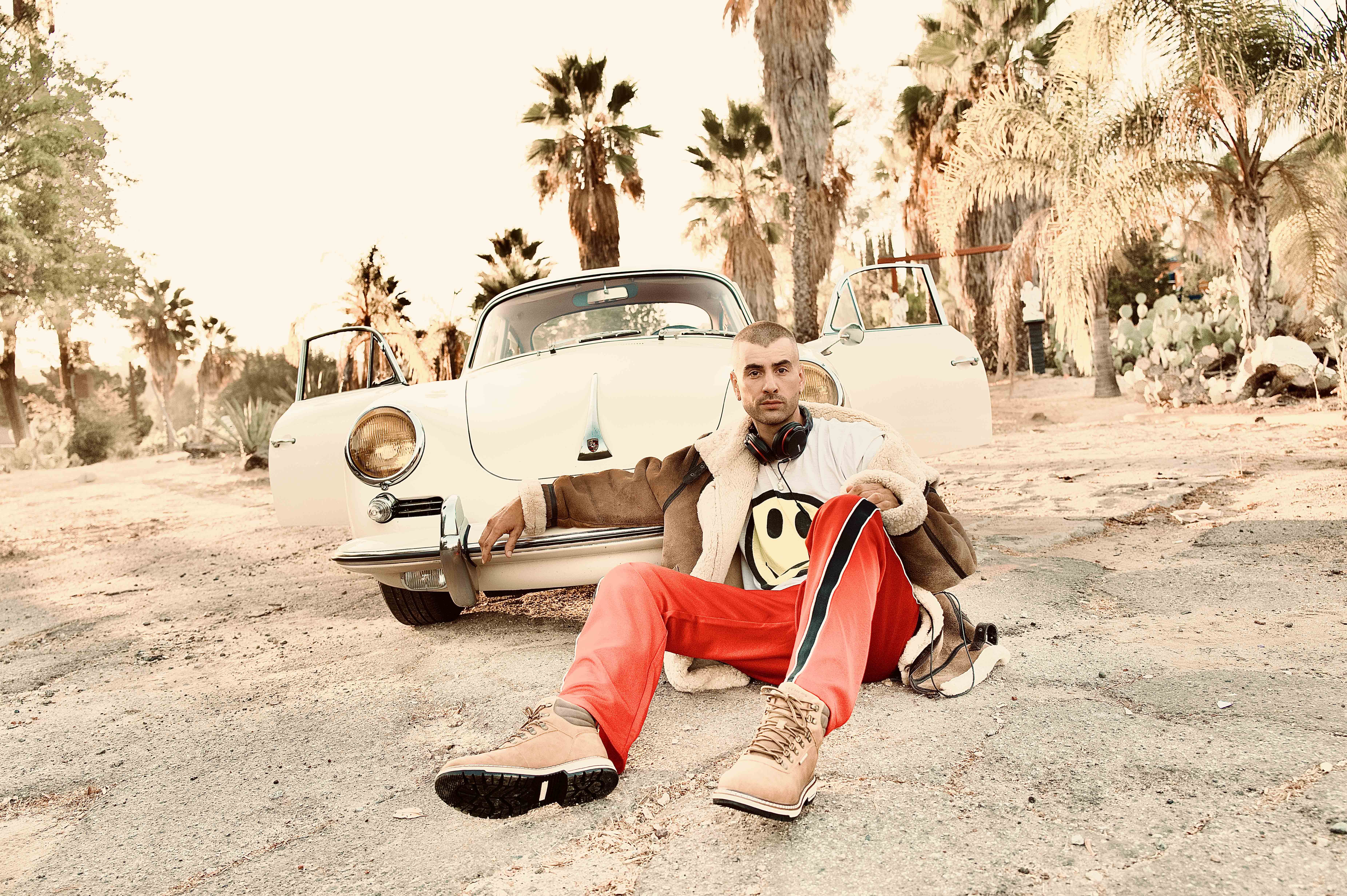 Adult male sits in front of an off-white vintage car, parked on a dirt road with palm trees and desert plants in the background.