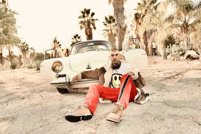 Adult male sits in front of an off-white vintage car, parked on a dirt road with palm trees and desert plants in the background.