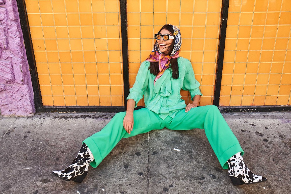 A cheerful young woman poses in stylish green outfit with headscarf and black and white boots against a yellow tiled wall outdoors.