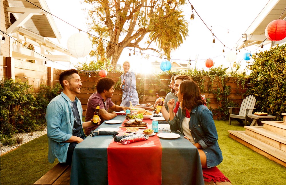 Group of friends enjoying an outdoor meal together at a table in a backyard, with string lights and colorful lanterns overhead.