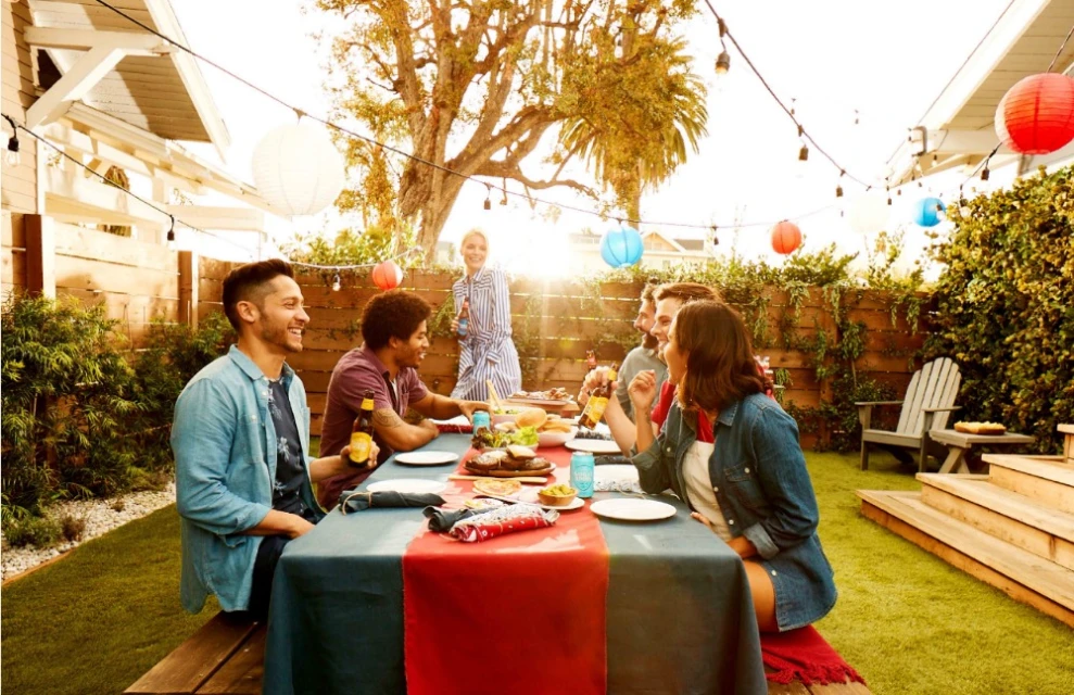 Group of friends enjoying an outdoor meal together at a table in a backyard, with string lights and colorful lanterns overhead.