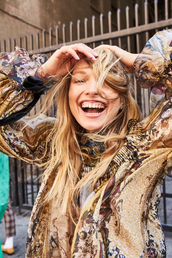 Smiling blonde woman with her hands in her hair wearing a patterned floral top outdoors in Los Angeles.