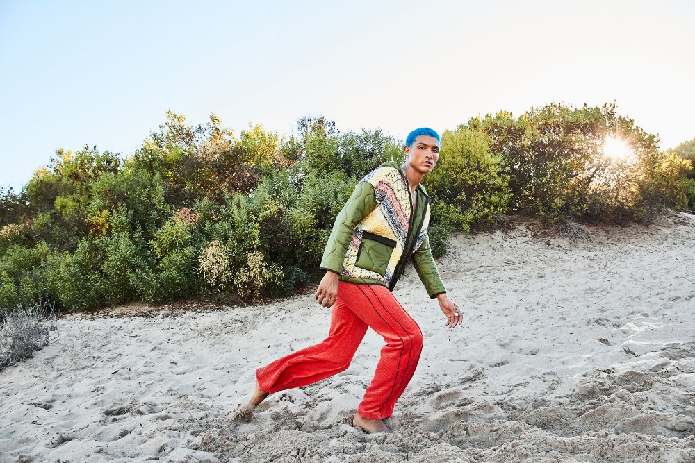 A person with blue hair wearing a quilted jacket and red pants walks up a sand dune with green shrubs in the background under a blue sky.