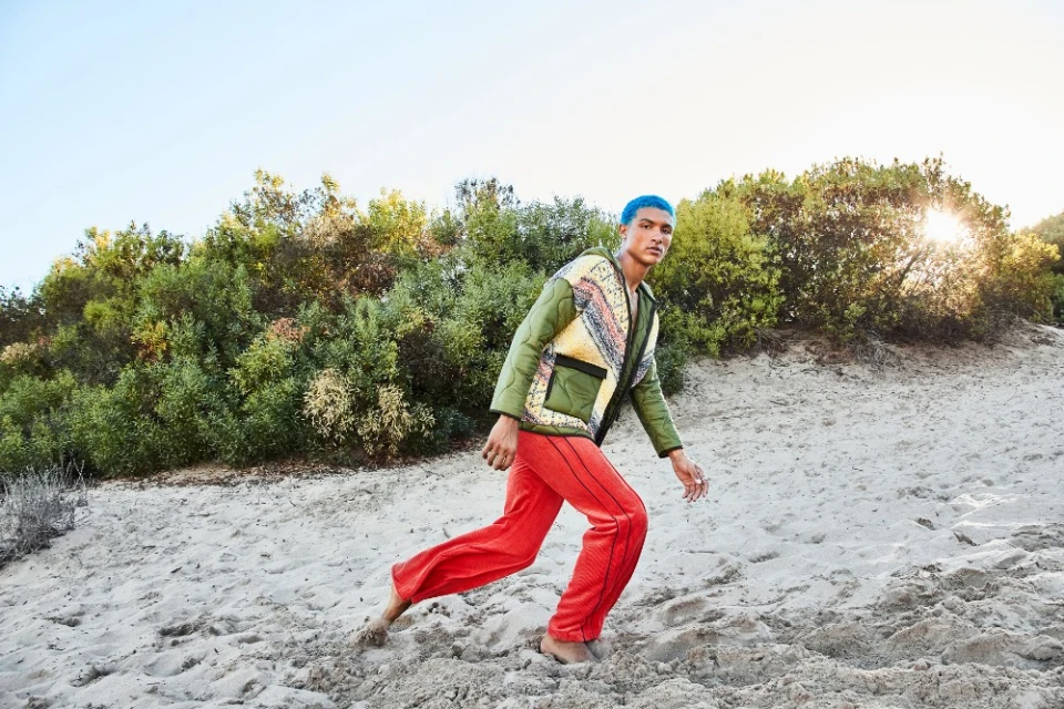 A person with blue hair wearing a quilted jacket and red pants walks up a sand dune with green shrubs in the background under a blue sky.
