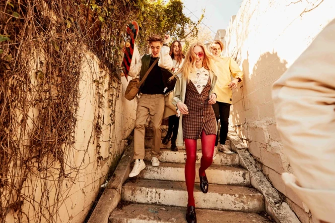 A group of young adults walks down sunlit stairs outdoors, showcasing casual fashion, carrying a skateboard, and enjoying a day out.
