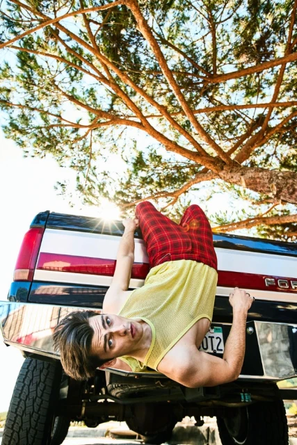 A young adult male hangs upside down from the back of a pickup truck, wearing red plaid pants, yellow shirt, and sunny outdoor lighting.