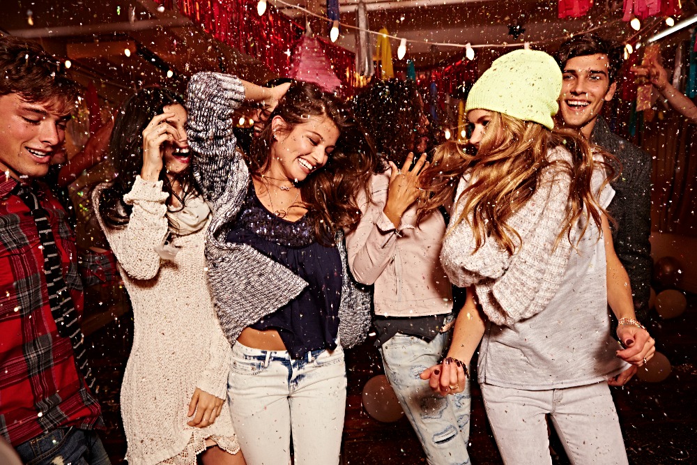 Group of six young adults celebrating with confetti indoors, dressed in casual clothes, and laughing in a candid, lively party setting.