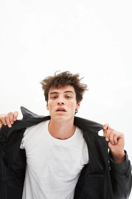 Portrait of a young adult male with curly brown hair, wearing a white t-shirt and partially wearing a black leather jacket against a white background.